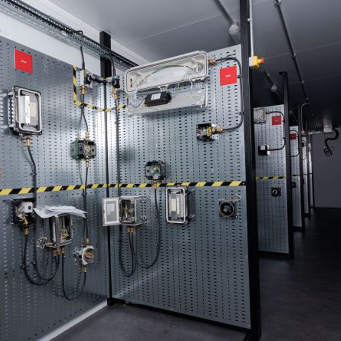 Industrial control room with various electrical panels mounted on perforated metal walls, featuring wiring, connectors, and caution tape. The room has a narrow walkway and industrial lighting.