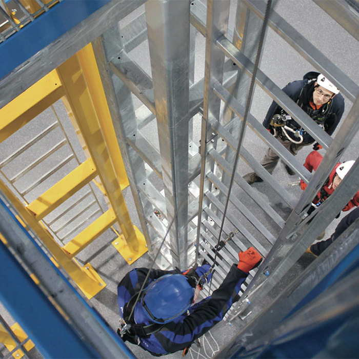 Workers equipped with safety gear are climbing a metallic industrial structure with ladders and platforms.