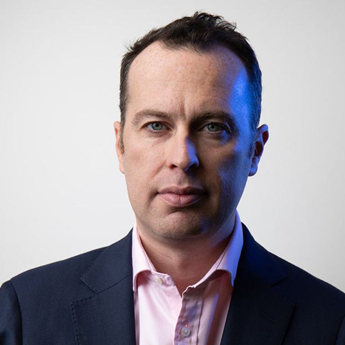 رجل ذو شعر داكن قصير، يرتدي بدلة زرقاء وقميصاً وردياً، ينظر مباشرة إلى الكاميرا على خلفية بيضاء عادية.