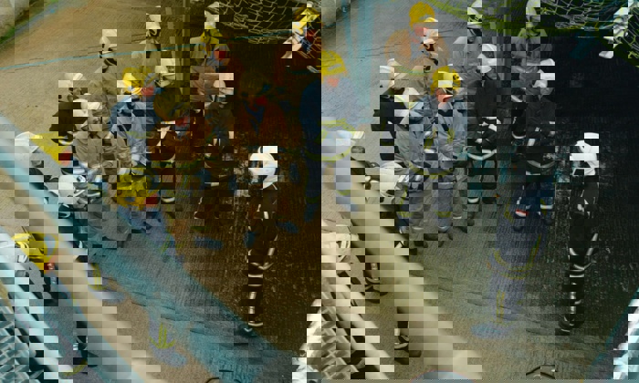 Un grupo de bomberos ataviados con cascos amarillos y equipo de extinción de incendios permanecen en formación, escuchando a otro bombero con casco blanco que les da instrucciones.