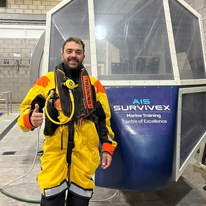 A man in a yellow and orange survival suit stands indoors, giving a thumbs-up. He is in front of a training simulator labeled "AIS Survivex Marine Training Centre of Excellence.