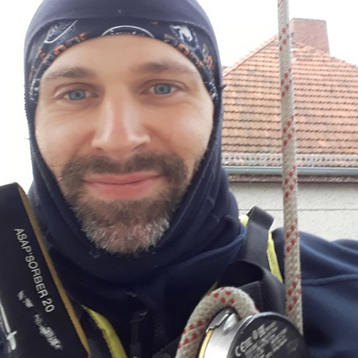 A person in outdoor gear, including a harness and ropes, smiles at the camera with a building and rooftop in the background.