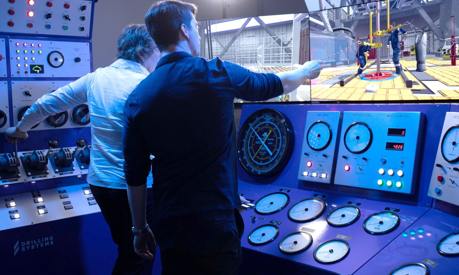 يقوم شخصان بتشغيل لوحة تحكم في غرفة محاكاة الحفر، مع وجود مقاييس وشاشات تعرض أنشطة الحفر في الوقت الفعلي.