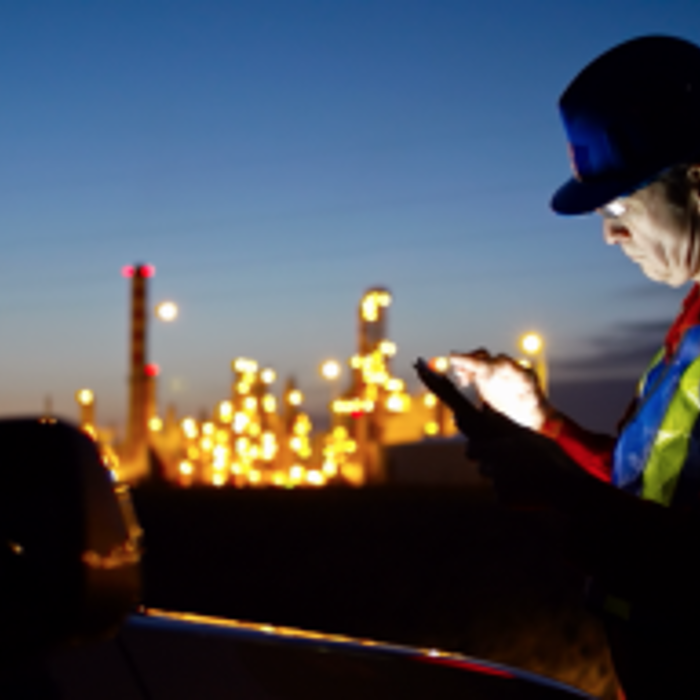 A worker in a hard hat and safety vest uses a tablet in a dimly lit industrial setting with bright lights in the background.