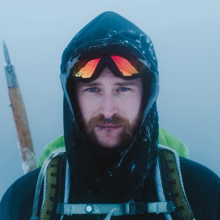 A person with facial piercings, sunglasses, and ice-covered clothing stands outdoors in snowy, foggy conditions. A hiking pole is attached to their backpack.