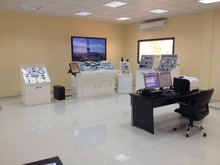 Uma sala de controlo com vários painéis de controlo, uma secretária com monitores de computador, vários manómetros e um grande ecrã na parede com a imagem de uma instalação industrial.