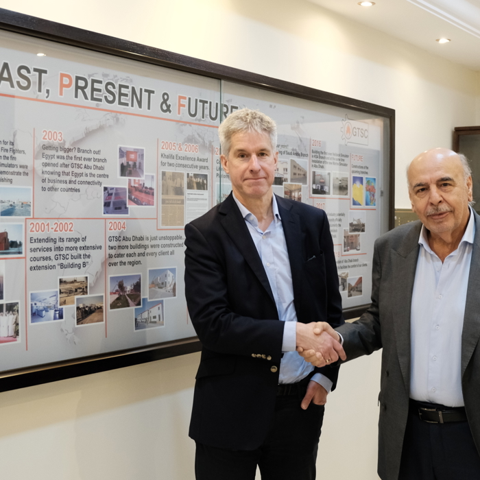 Two men shaking hands in front of a timeline display board labeled "GTSC Past, Present & Future" in an office, setting the stage for future training initiatives.