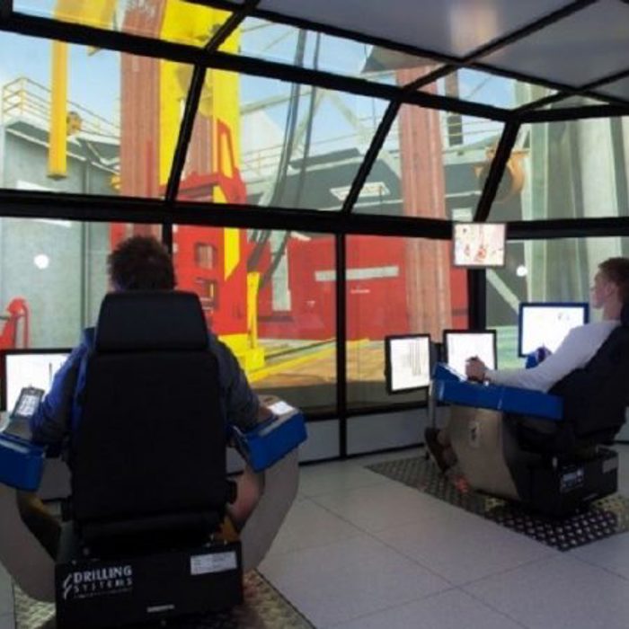 Two people operating simulators in a room with large screen displays showing an industrial environment, possibly for training purposes.