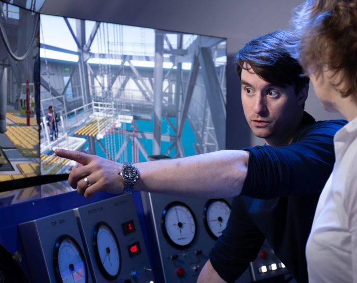 شخص يشير إلى شاشة تعرض إعدادات صناعية أثناء شرح شيء ما لشخص آخر. تظهر العدادات والمقاييس على لوحة تحكم في المقدمة.