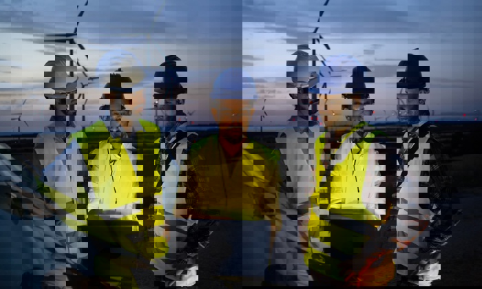 Três trabalhadores com equipamento de segurança e capacetes recebem formação enquanto examinam um computador portátil perto de turbinas eólicas ao anoitecer.